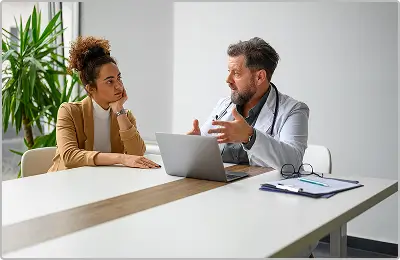 A doctor sits at a desk with a laptop, talking to a woman who listens attentively. The doctor gestures with his hands while the woman rests her chin on her hand. A clipboard and glasses are on the table.